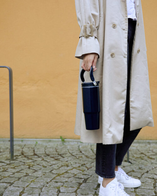 Nainen beigessä trenssissä kantaa vasemmassa kädessä mustaa termosmukia