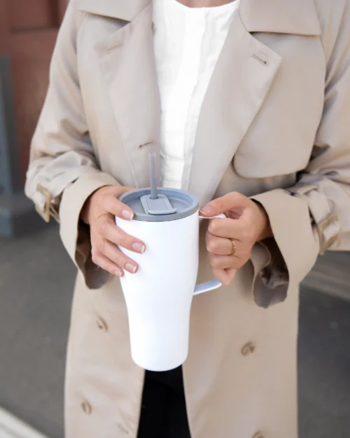 Nainen beigessä trenssissä pitää kädessään valkoista pillillistä termosmukia