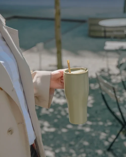 Nainen beigessä trenssissä pitää vasemmassa kädessään vihreää pillillistä termosmukia